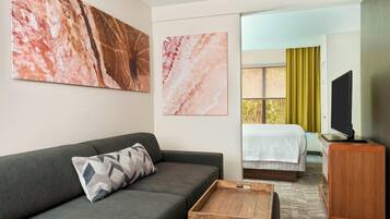 Une chambre d’hôtel moderne avec un canapé gris, une table d’appoint en bois, un lit avec une couette blanche, un téléviseur posé sur un meuble en bois, et deux grandes œuvres d’art en marbre accrochées au mur.