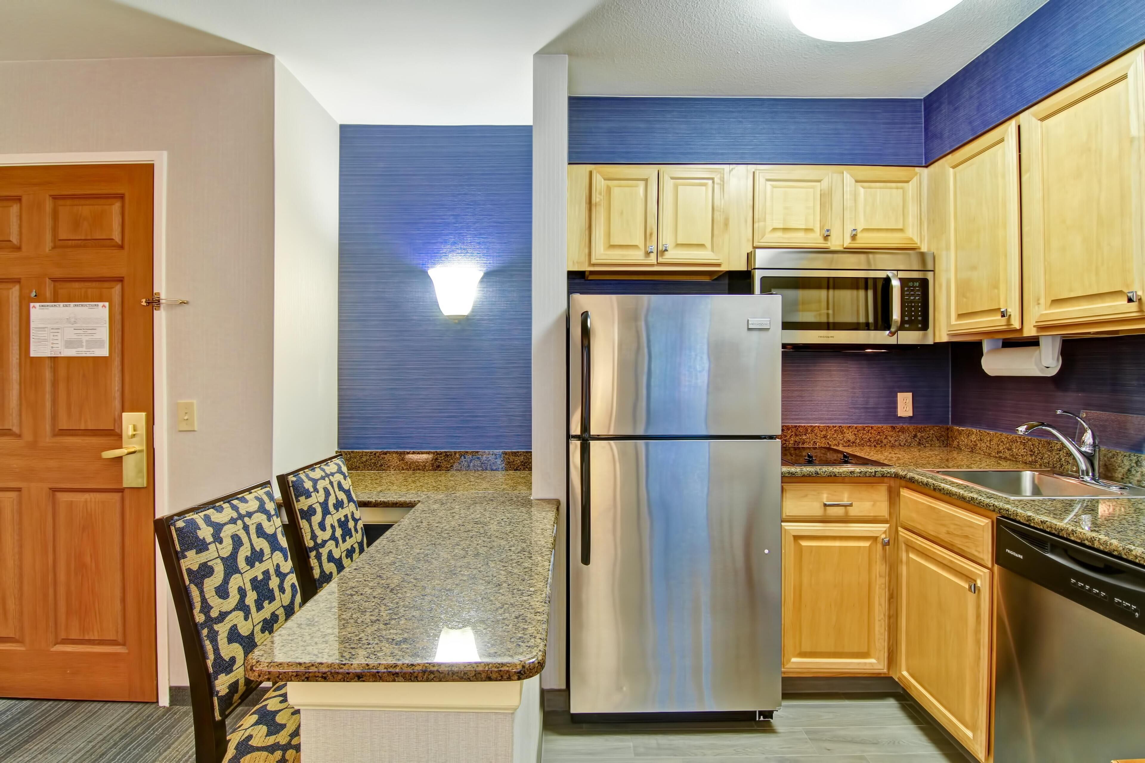 A compact kitchen with a refrigerator, microwave, and sink. There is a dining area with a table and chairs. The kitchen has wooden cabinets and a blue accent wall.