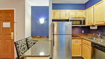 A compact kitchen with a refrigerator, microwave, and sink. There is a dining area with a table and chairs. The kitchen has wooden cabinets and a blue accent wall.