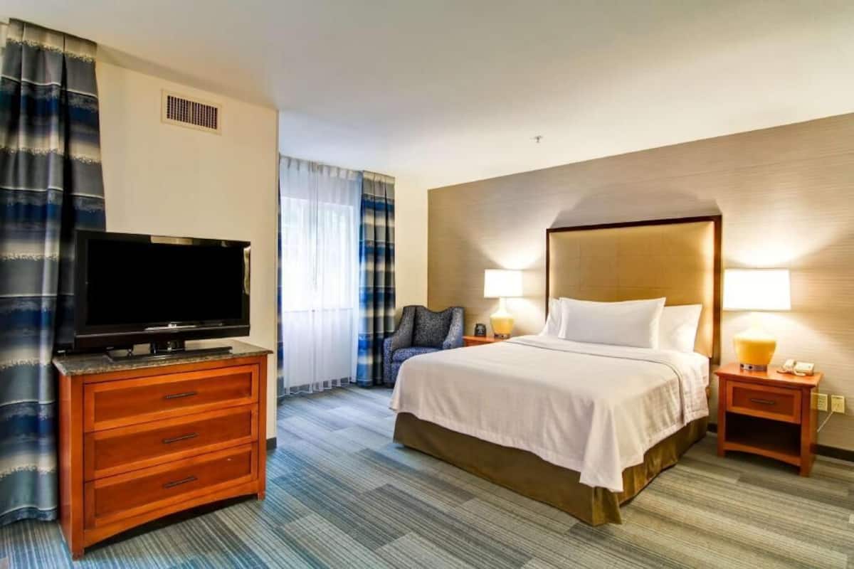 Ein Hotelzimmer mit einem Bett, einem Fernseher auf einer Holzkommode, einem Nachttisch mit Lampe und einem Sessel.