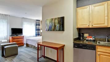 Ein Hotelzimmer mit einem Bett, einem Fernseher, einem Holztisch, einer Küchenzeile inklusive Geschirrspüler und einem Bild an der Wand.