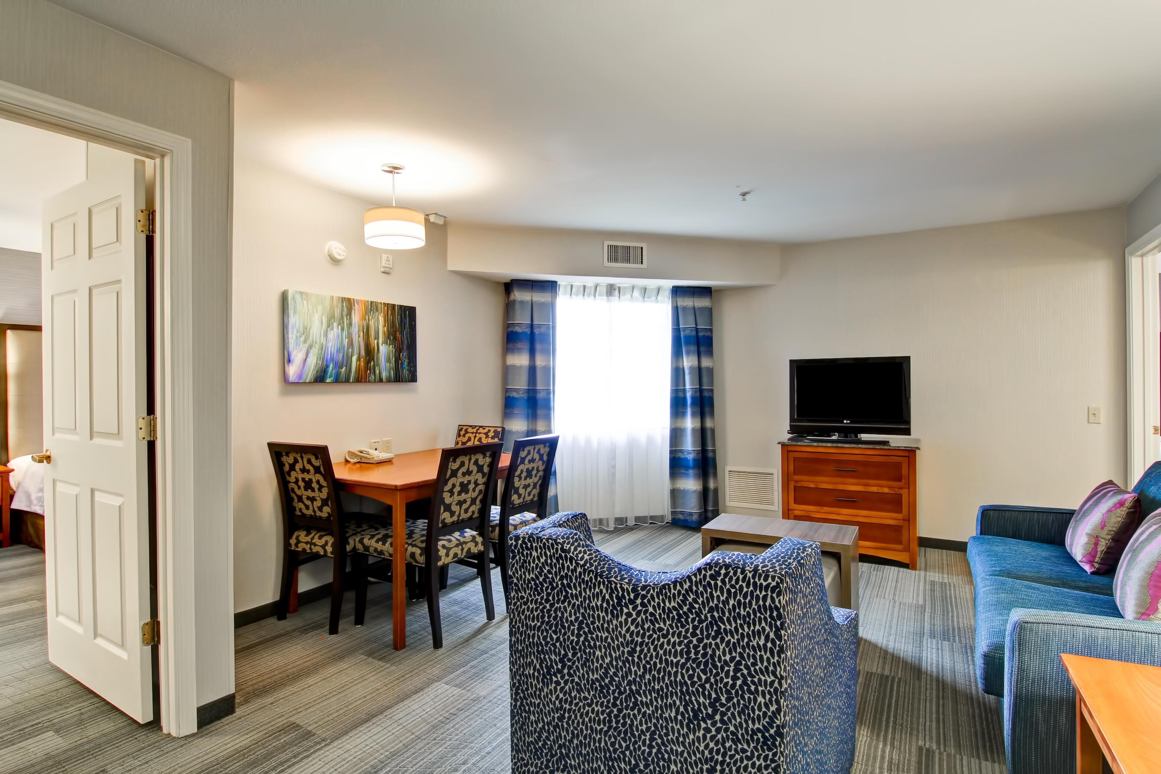 Habitación de hotel con comedor, sofá, televisor y un cuadro en la pared.