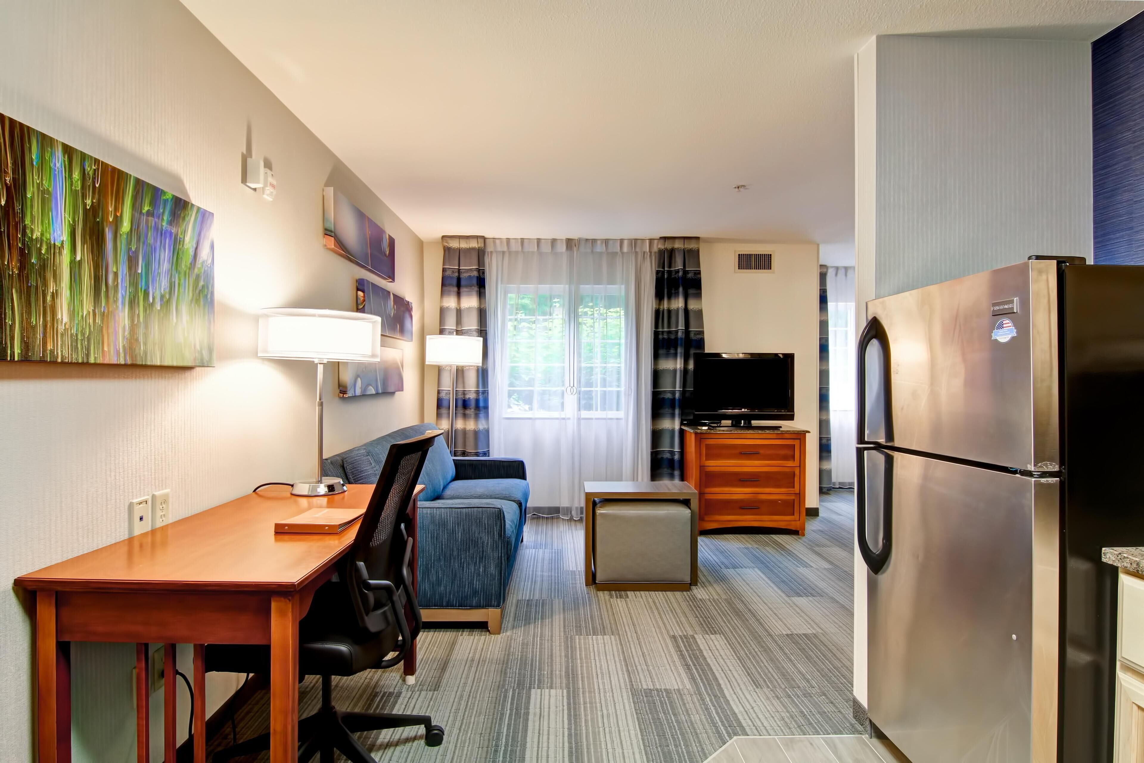 A compact hotel room with a kitchenette, a sofa, a desk, and a TV.