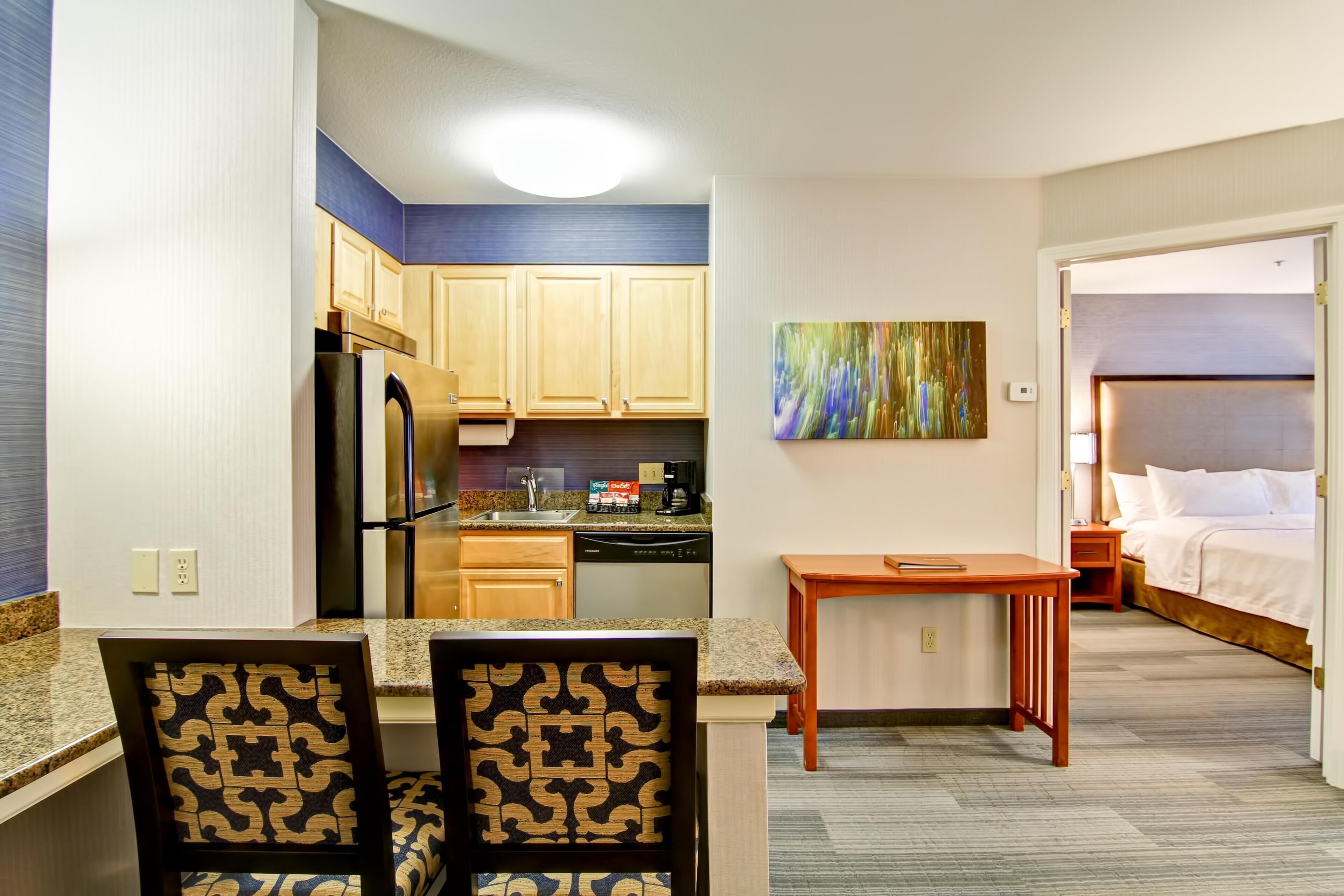 A compact hotel room with a kitchenette, a dining area with patterned chairs, a small table, and a bed with a headboard.