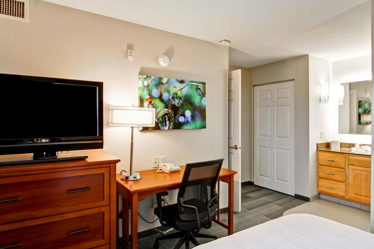 Ein Hotelzimmer mit einem Bett, einem Schreibtisch mit Computer, einem Fernseher, einer Lampe, einem Stuhl und einem Bild an der Wand.