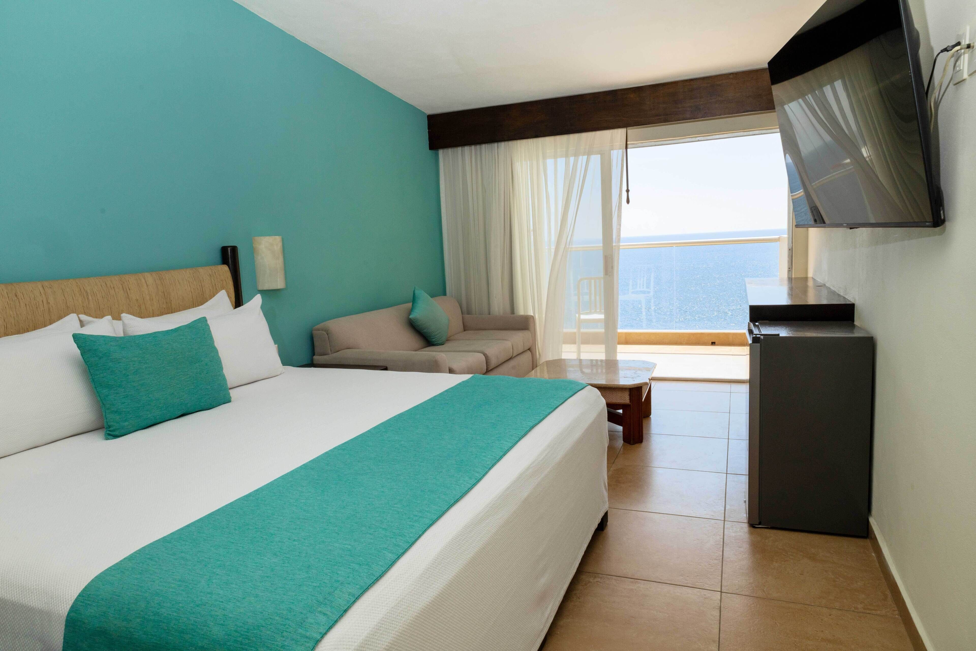 Quarto de hotel com cama, sofá, televisão e varanda com vista.