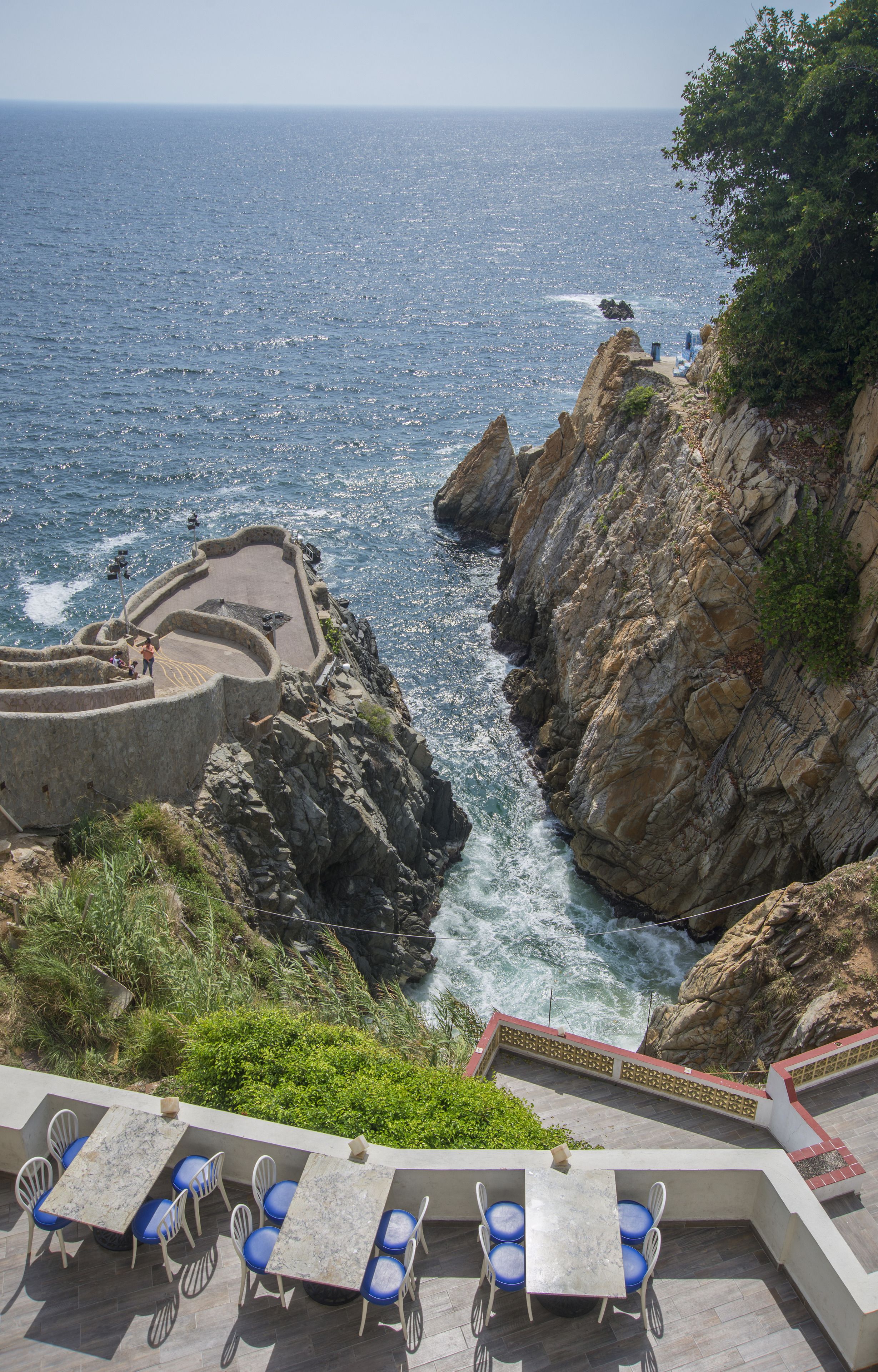 A terrace with outdoor seating overlooking the sea, a rocky cliff, and a clear view of the ocean.