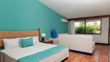 Una habitación de hotel con una cama, una mesita de noche, una jarra de agua azul, un sofá blanco y una ventana con vistas a edificios y árboles.