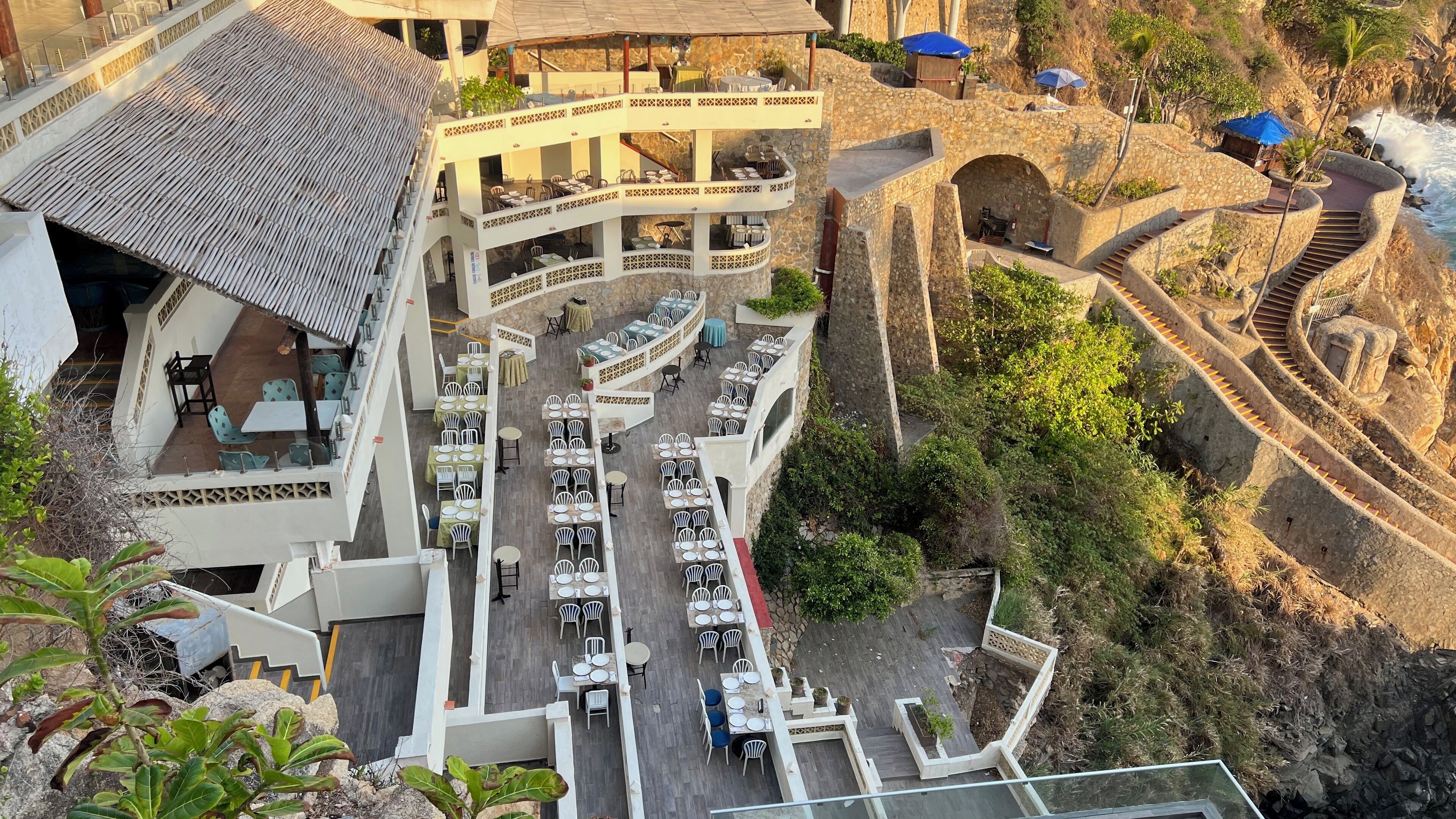 A multi-level outdoor restaurant with terraces and dining areas, surrounded by stone walls and a coastal view.
