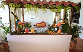 Zona de bar con temática tropical, con un centro de mesa con forma de guitarra, rodeada de plantas en macetas y diversas frutas.