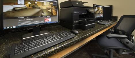 A hotel reception desk with two monitors, a desktop computer, a printer, and a guest chair.