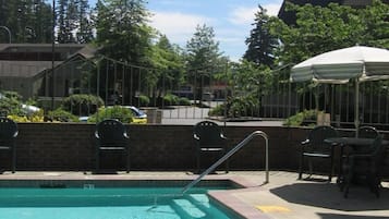 Une piscine avec une terrasse carrelée, entourée de chaises et de parasols, avec des arbres et des bâtiments en arrière-plan.