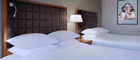 A hotel room with two beds, a headboard with a padded design, a bedside table with a lamp, and a framed picture on the wall.