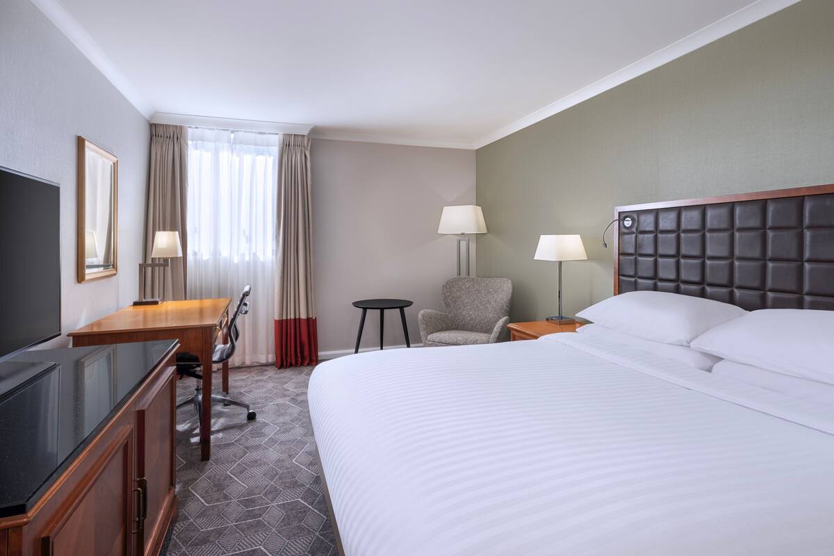 A hotel room with a large bed, a desk with a TV, a chair, two bedside lamps, and a window with curtains.