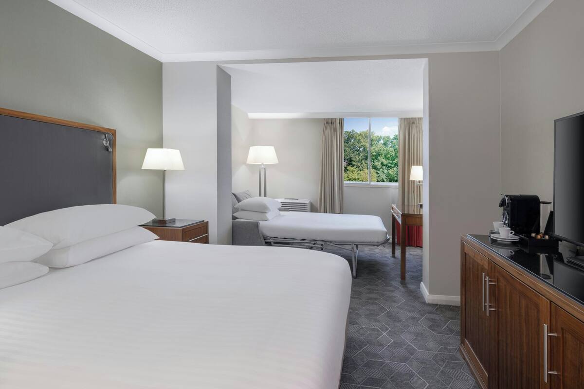 A hotel room with a large bed, a smaller bed, a desk with a lamp, a TV, and a view of greenery through the window.