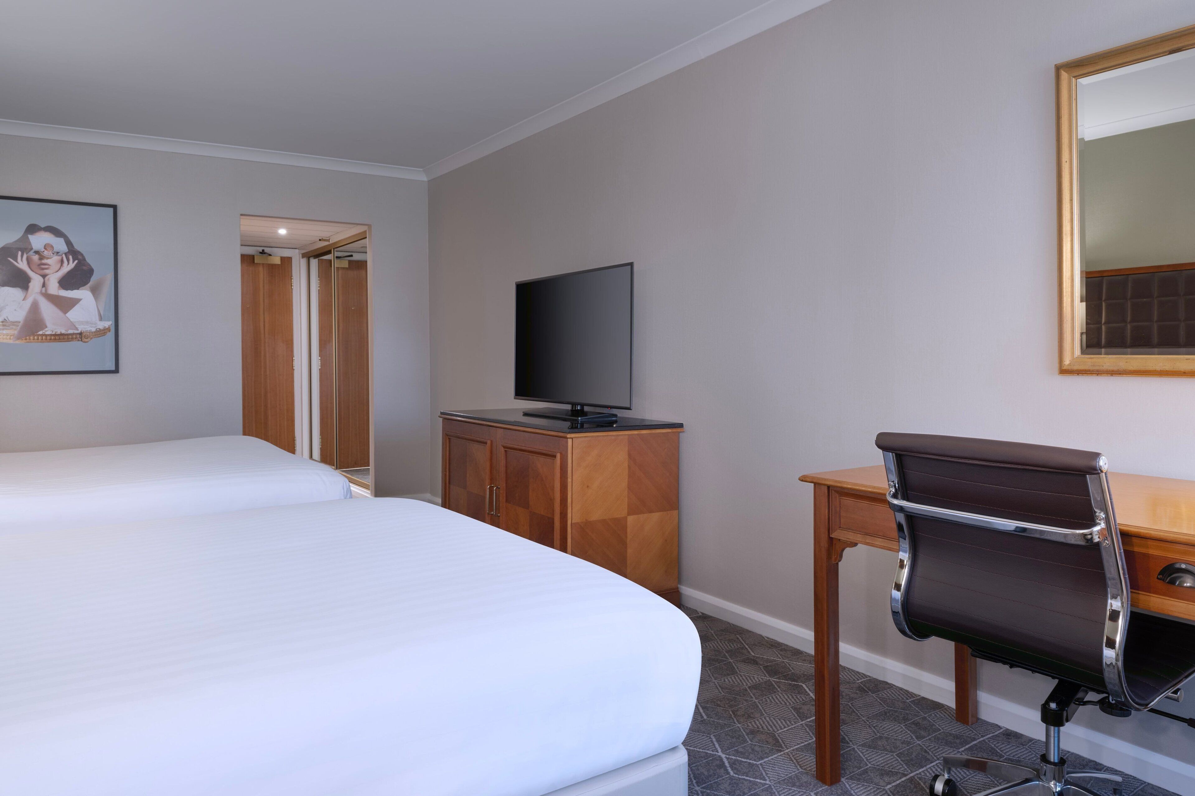 Une chambre d’hôtel avec deux lits, un bureau en bois, une chaise, une télévision et un miroir.