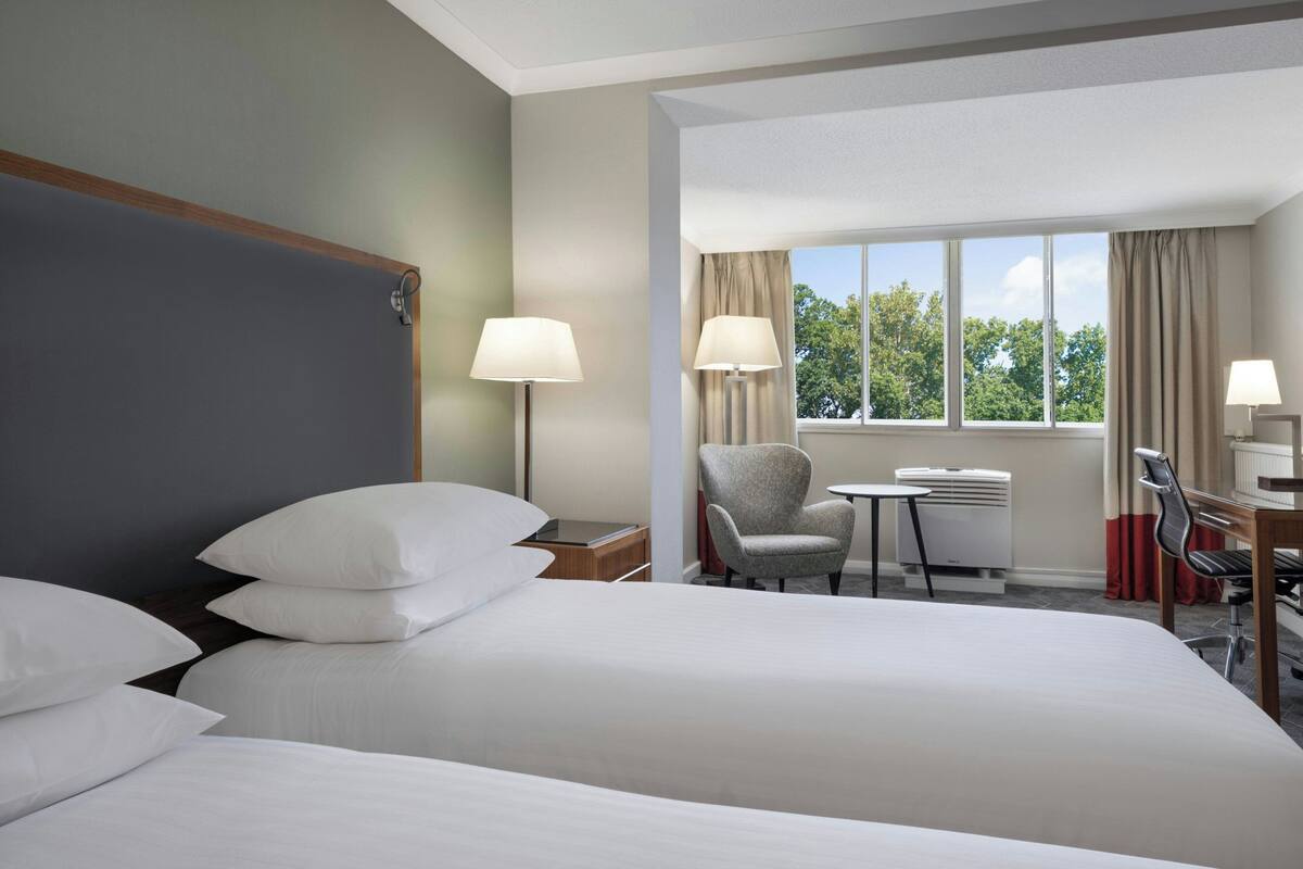 A hotel room with a large bed, two bedside lamps, a chair, a small table, and a window with curtains.