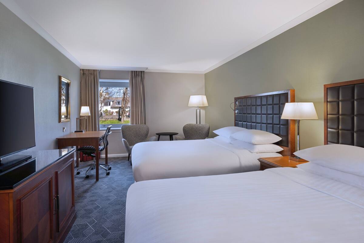 A hotel room with two beds, a desk, a chair, a TV, and a window with curtains.