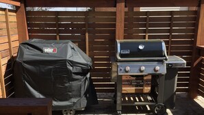Un espace patio équipé d’un barbecue Weber et d’un chariot de service couvert pour le barbecue, le tout adossé à une clôture en bois et posé sur un sol en briques.