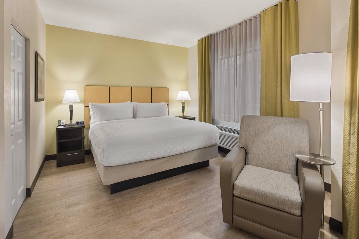Une chambre d’hôtel comprenant un lit, une chaise, deux tables de chevet avec des lampes et une fenêtre avec des rideaux.