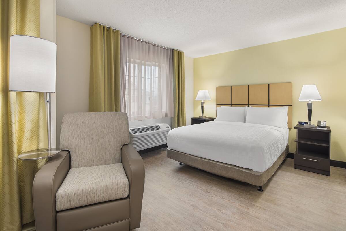 Une chambre d’hôtel avec un lit, une chaise, une table de chevet, une lampe et une fenêtre avec des rideaux.