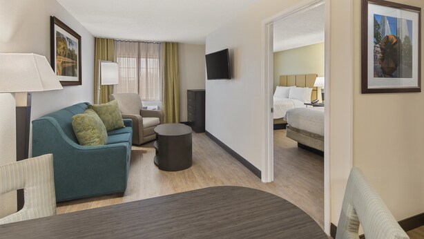 Une chambre d’hôtel avec un canapé, un fauteuil et un lit. Un téléviseur à écran plat est fixé au mur, ainsi qu’une lampe et un cadre photo.