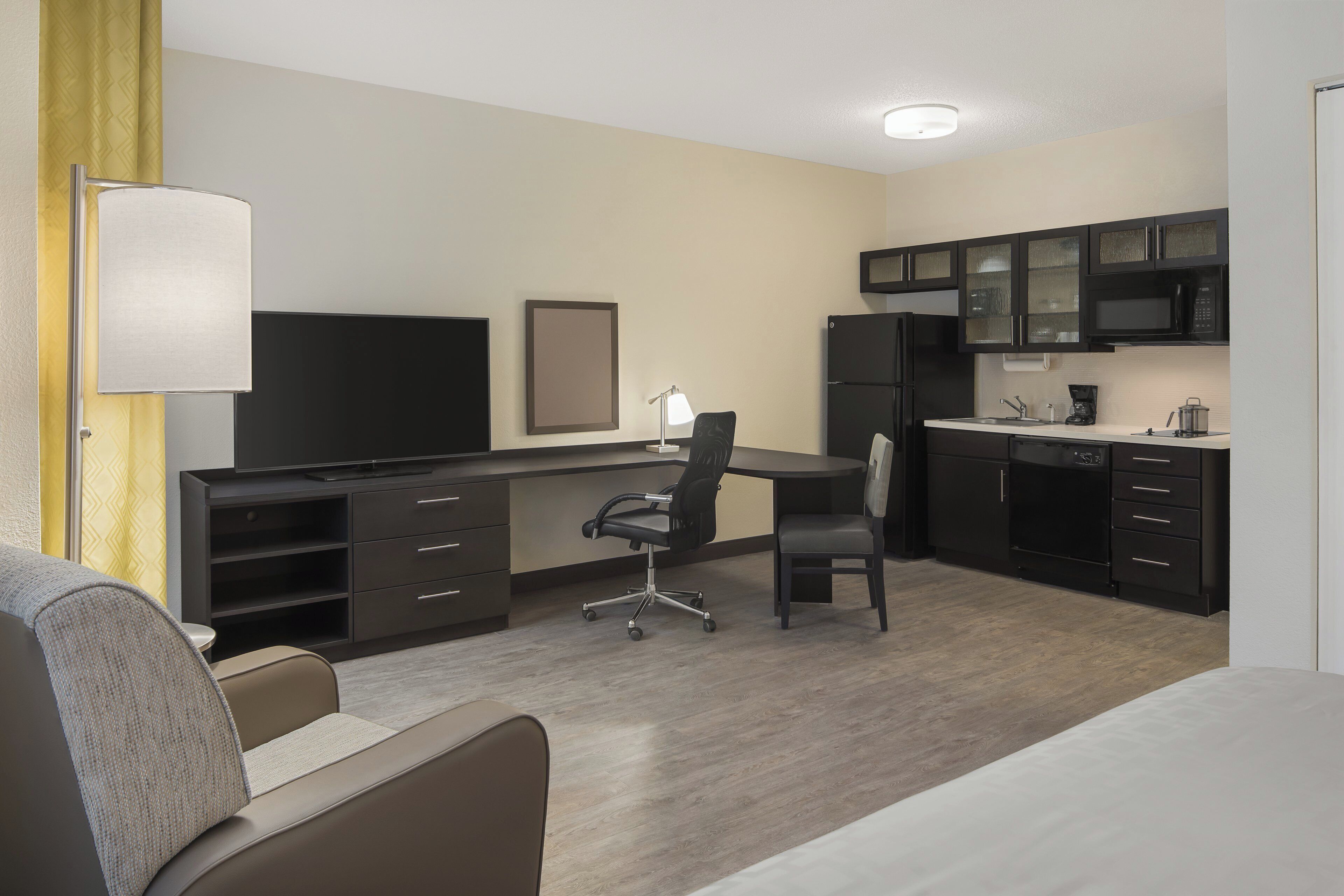 A hotel room with a bed, a desk, a chair, a television, a kitchen area with a microwave, and a refrigerator.