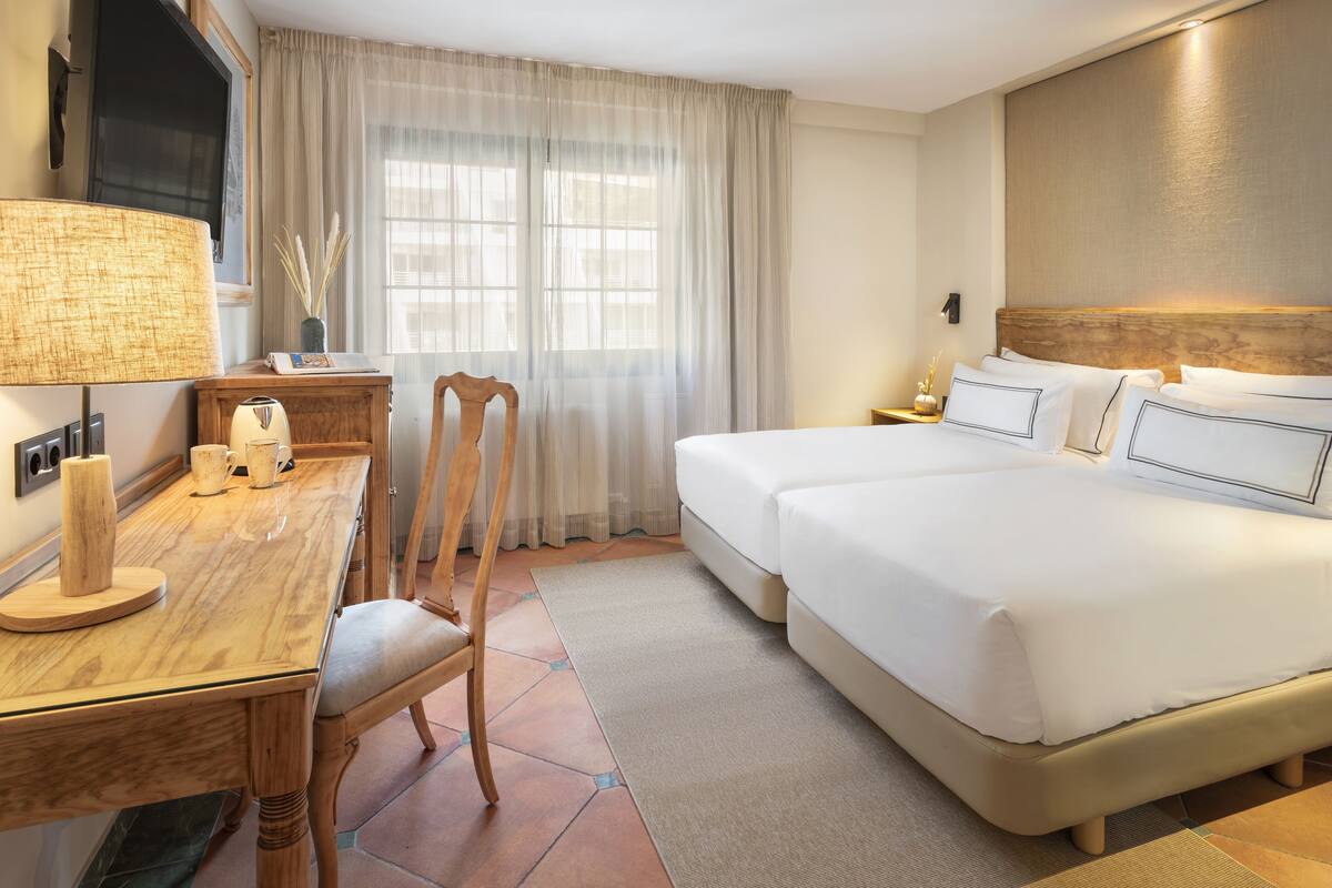 Habitación de hotel con un escritorio de madera, dos camas, un televisor y un ventanal grande con cortinas.