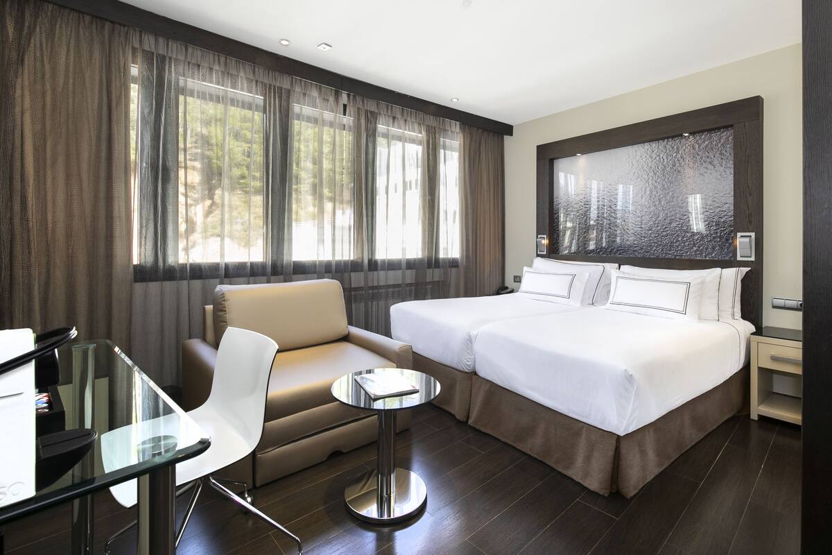 Una habitación de hotel moderna con una cama grande, un sofá, una mesita y un ventanal con cortinas.