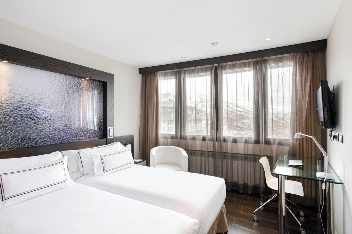 Habitación de hotel con dos camas, un escritorio, una silla, un televisor y un ventanal con cortinas.