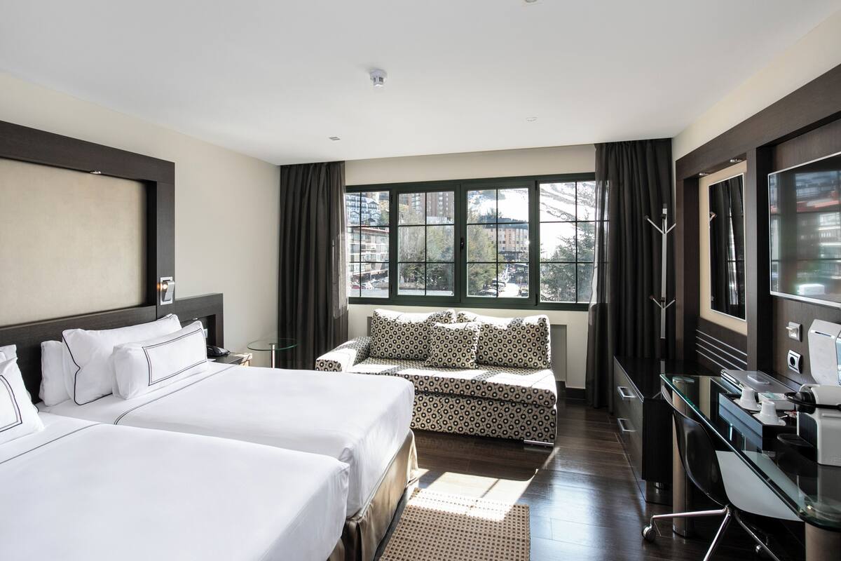 Habitación de hotel con dos camas, un sofá, un escritorio con computadora y un ventanal con cortinas.