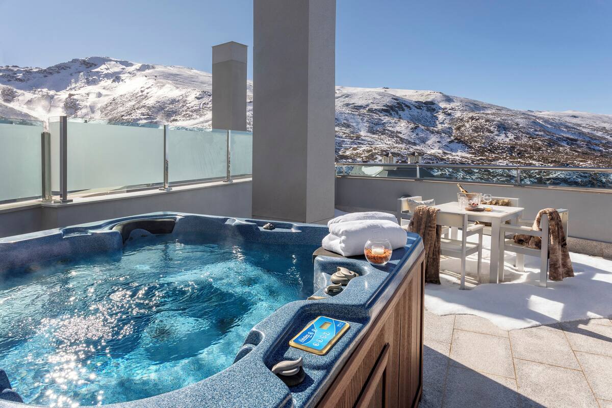Un jacuzzi con borde de madera, una mesa con sillas y vistas a montañas nevadas.
