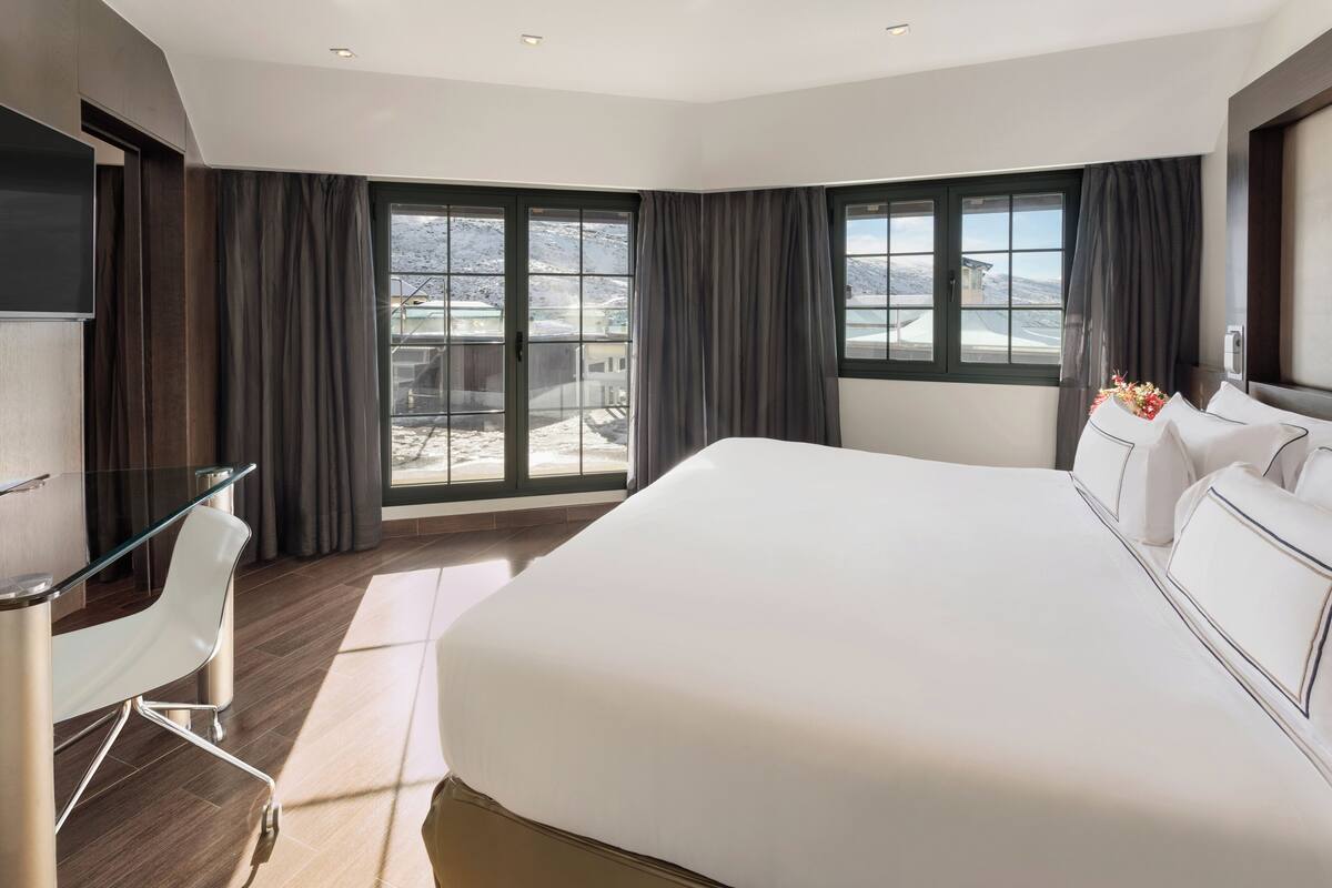 Una habitación de hotel moderna con una cama grande, un escritorio con silla y ventanas amplias con cortinas.