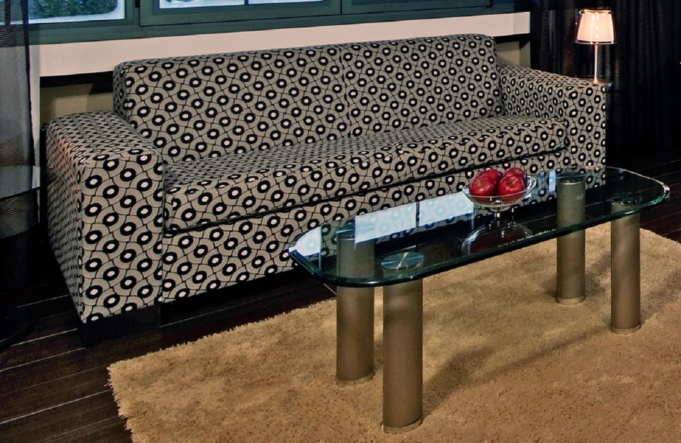 A patterned sofa, a glass coffee table with a bowl of fruit, and a lamp.