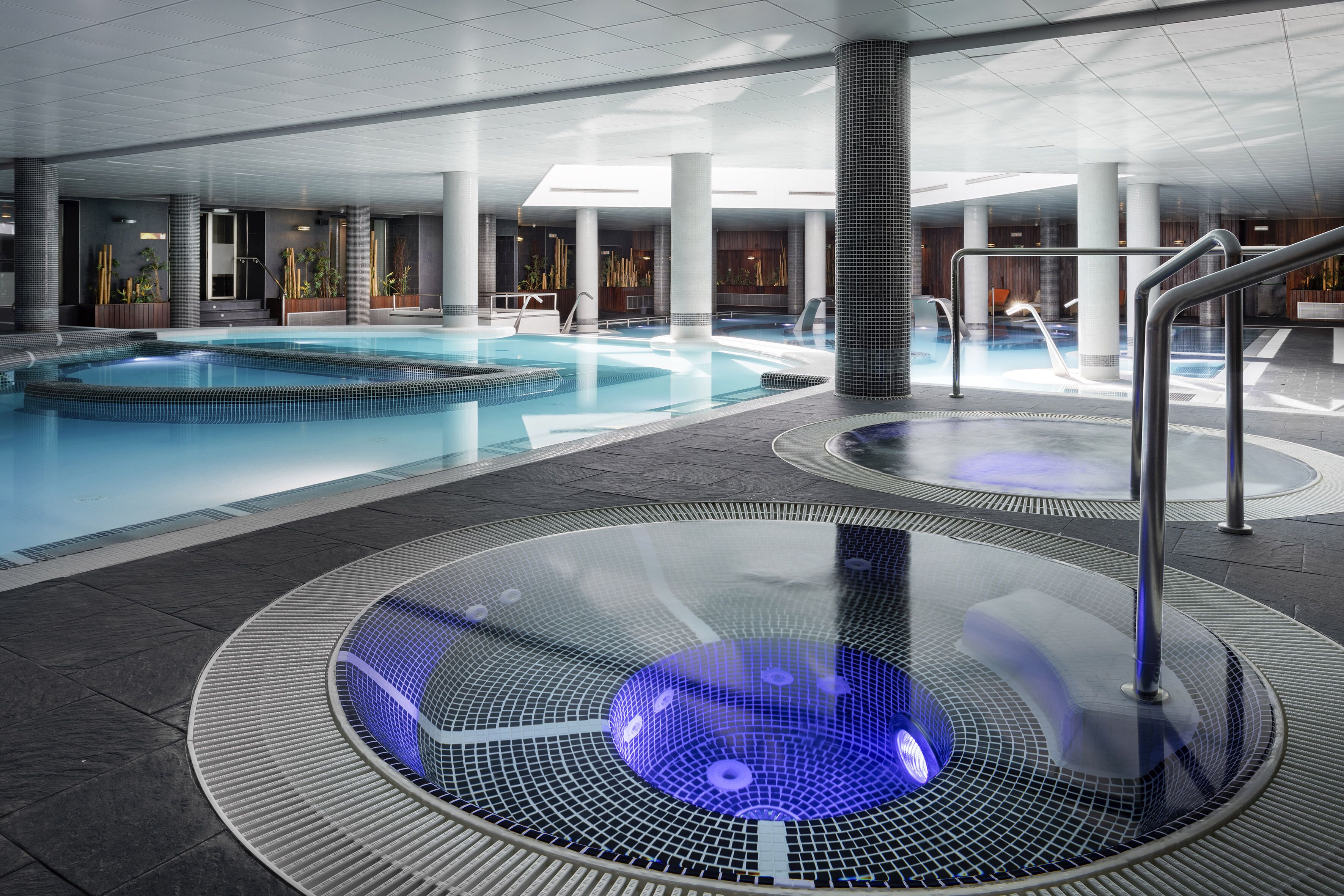 Ein moderner, überdachter Pool mit Whirlpool, umgeben von Säulen und einer Glasscheibe.