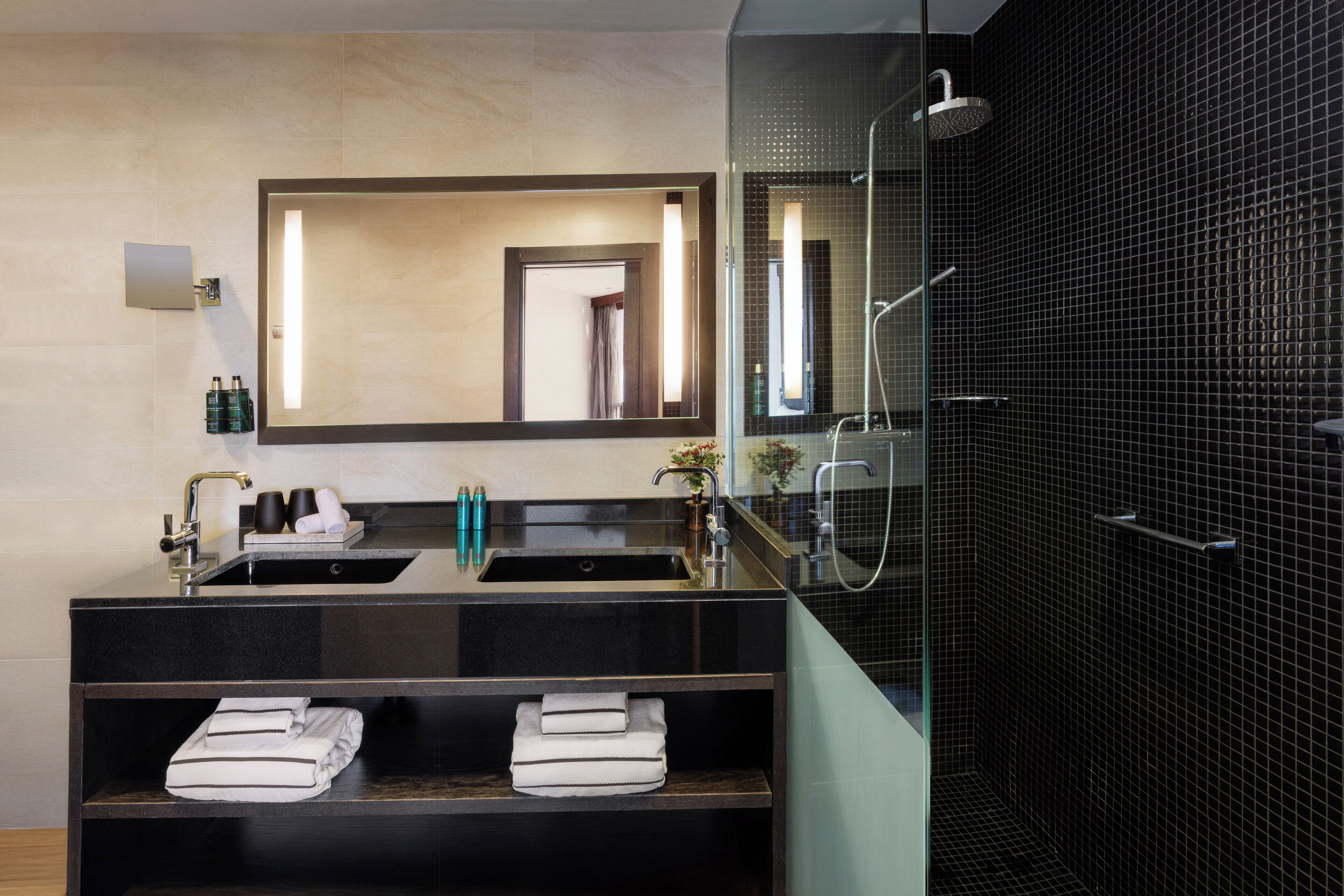 Une salle de bain moderne avec un meuble-lavabo à deux vasques, un grand miroir, une douche à parois de verre et des serviettes soigneusement pliées, rangées sur des étagères.