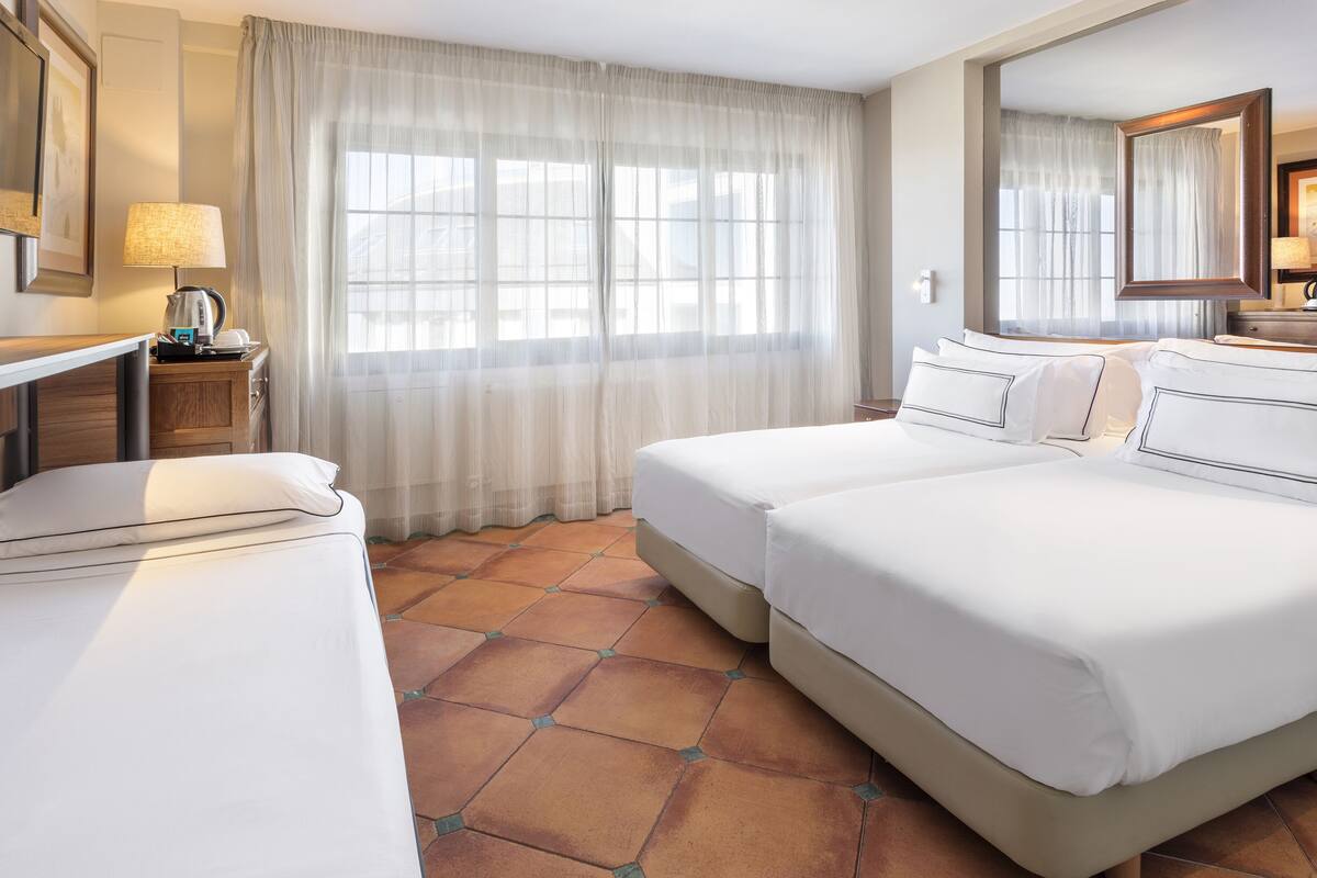 Habitación de hotel con dos camas, un ventanal grande con cortinas translúcidas, una mesita de noche con lámpara y piso de baldosas.