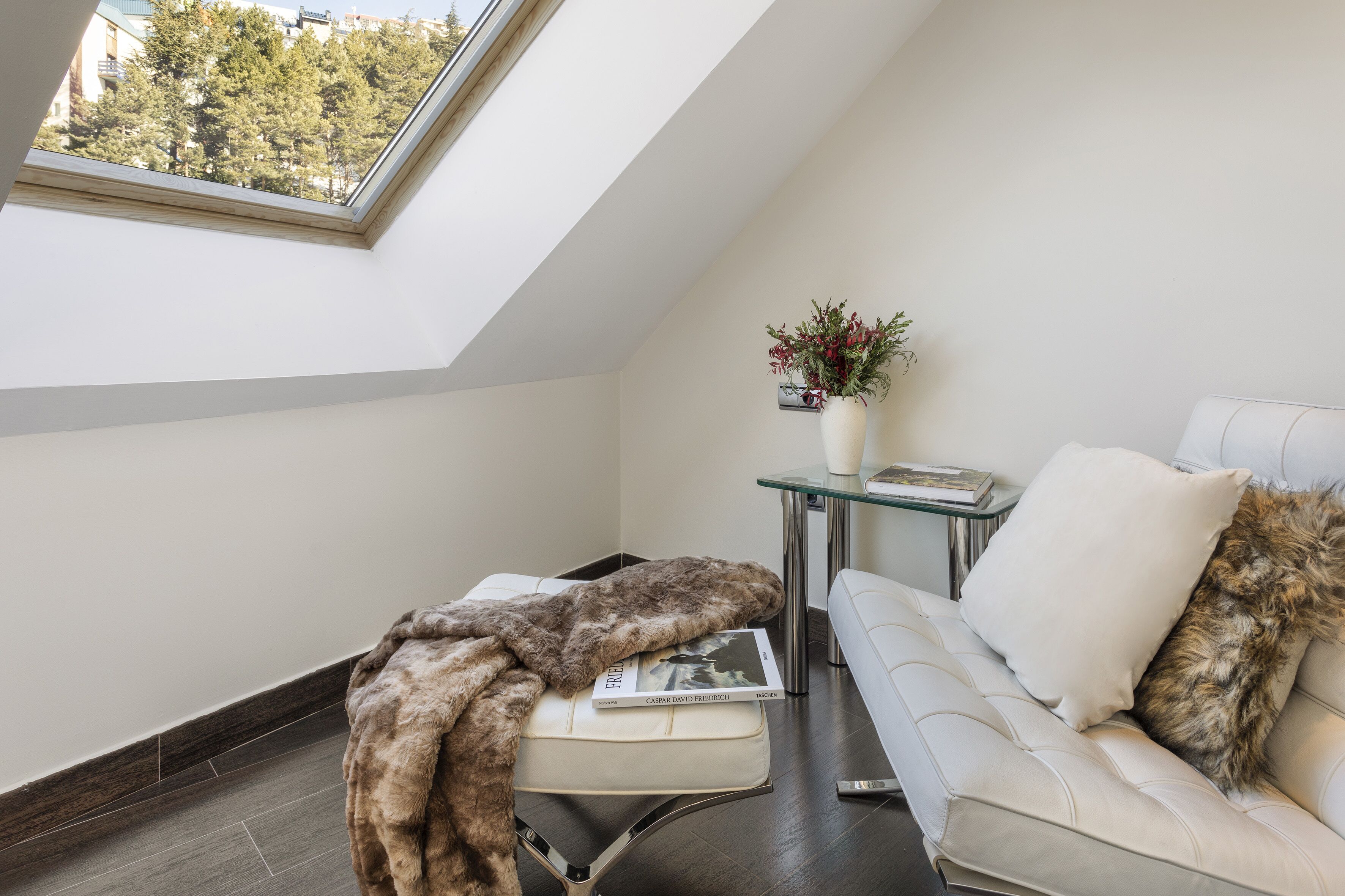 A modern loft bedroom with a skylight, a white sofa, a glass coffee table, and a decorative vase.