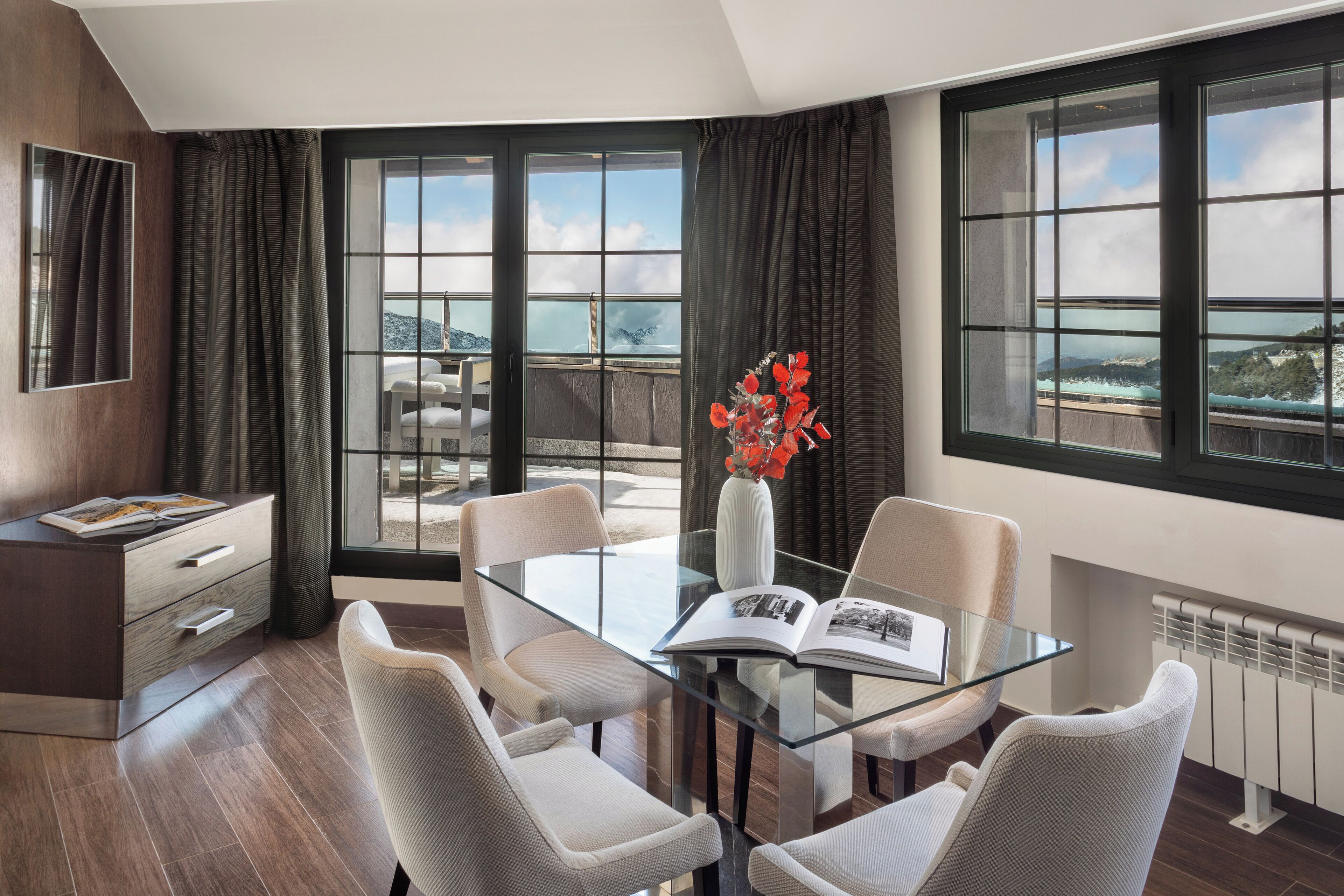 A modern room with a glass table, four chairs, a book, and a vase with flowers.