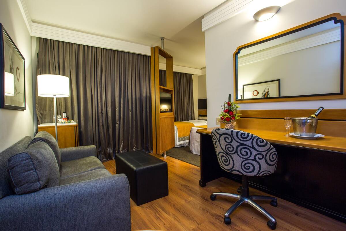 Habitación de hotel con un sofá gris, un otoman negro, un escritorio con una silla, un espejo grande y una cama con cabecera de madera.
