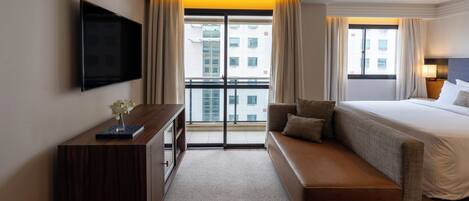 A hotel room with a large bed, a sofa, a TV mounted on the wall, and a balcony with curtains.