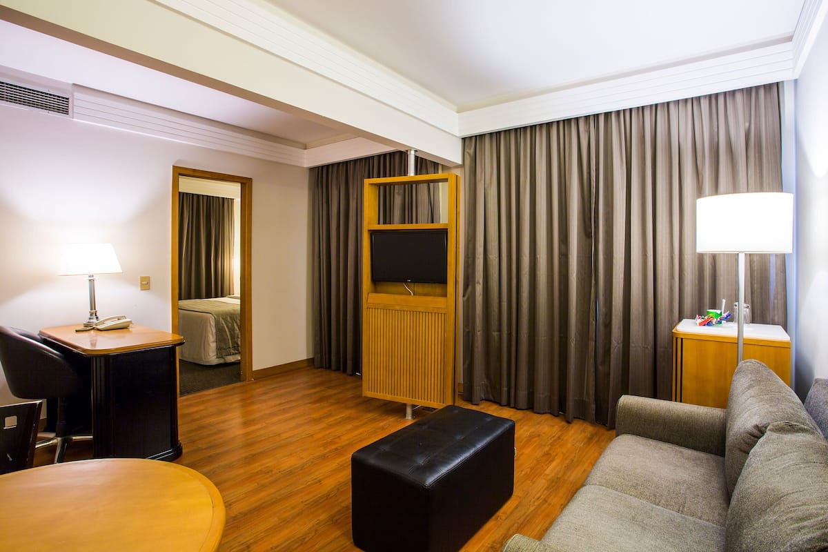 Habitación de hotel con piso de madera, sofá, una pequeña mesa con lámpara, un mueble para televisión y una cama con cortinas.
