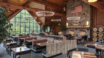 A spacious restaurant with high ceilings, wooden beams, and large windows offering a view of the outdoors. There are multiple seating areas, including booths and tables, and a bar area with a stone wall and American flag.