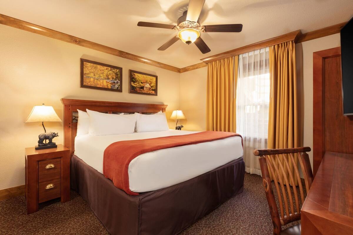 Une chambre d’hôtel avec un lit, deux lampes de chevet, un ventilateur de plafond, une table de chevet en bois, une chaise et deux tableaux encadrés au mur.