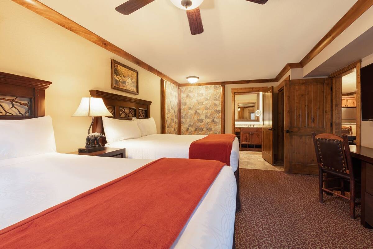 Une chambre d’hôtel avec deux lits, un bureau, une chaise, un ventilateur de plafond, un mur décoratif en pierre et une porte en bois.