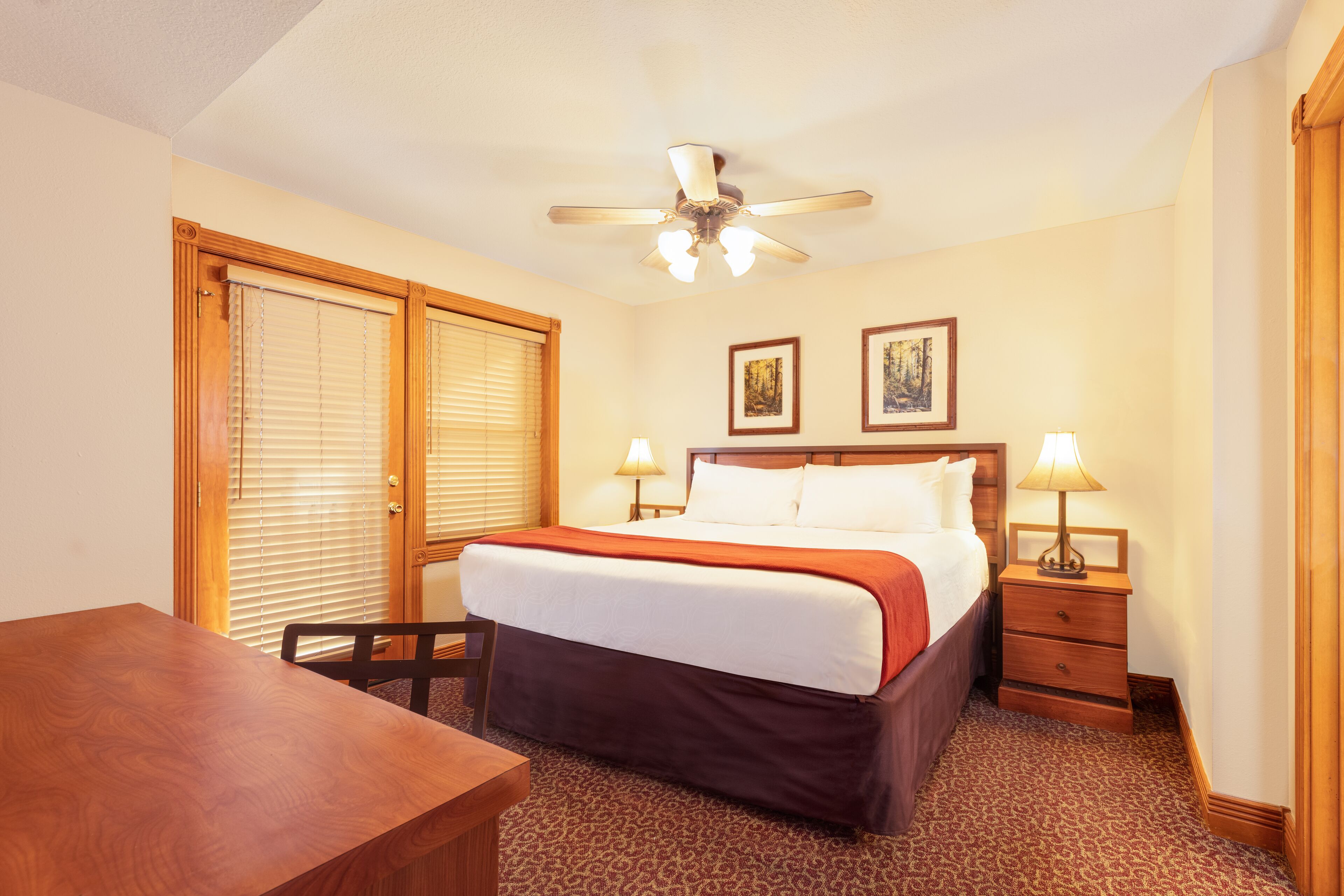 A hotel room with a bed, a desk, two bedside lamps, a ceiling fan, and two framed pictures on the wall.