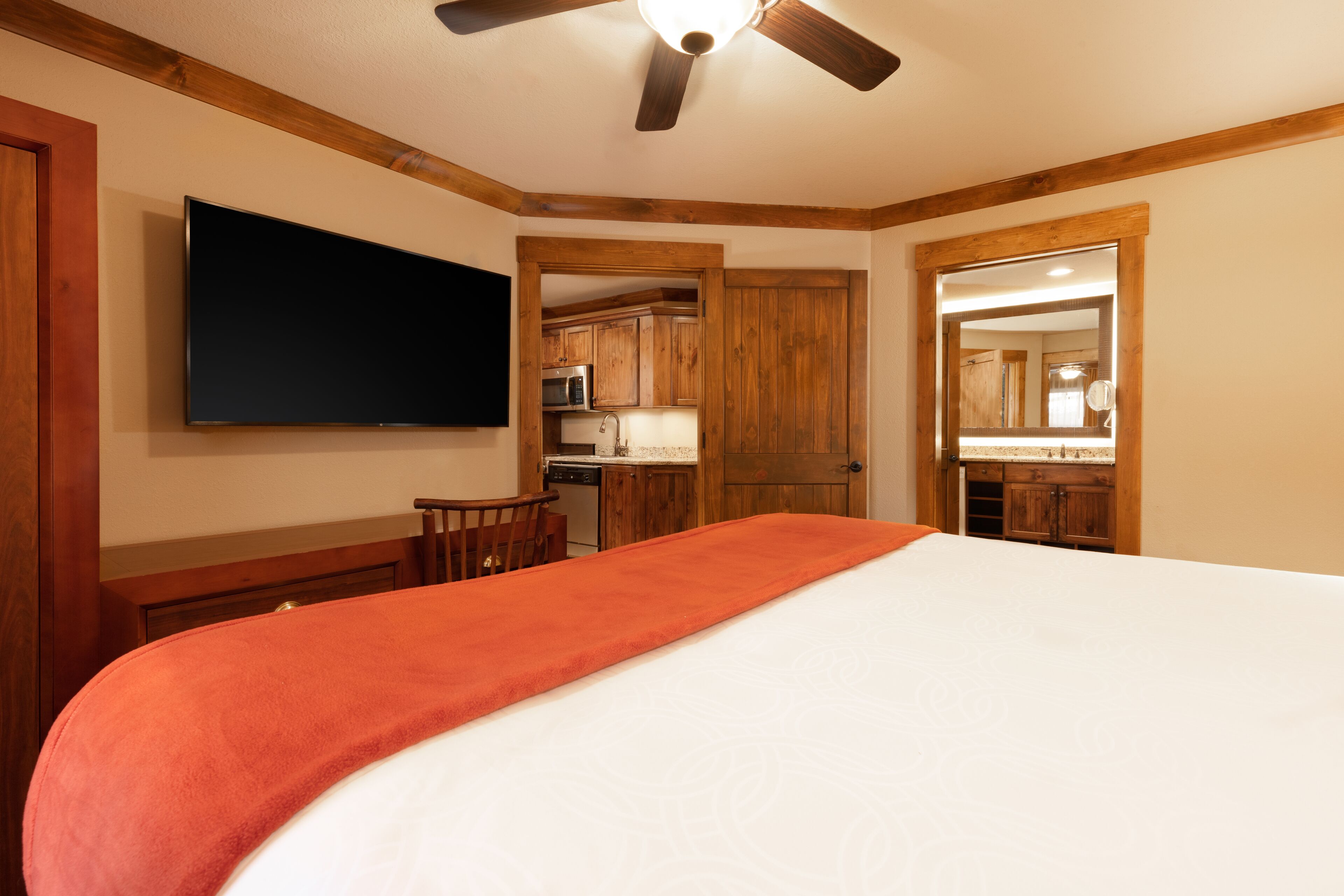 Une chambre avec un lit, un téléviseur fixé au mur, un ventilateur de plafond, des meubles en bois et un coin cuisine visible à travers une porte ouverte.