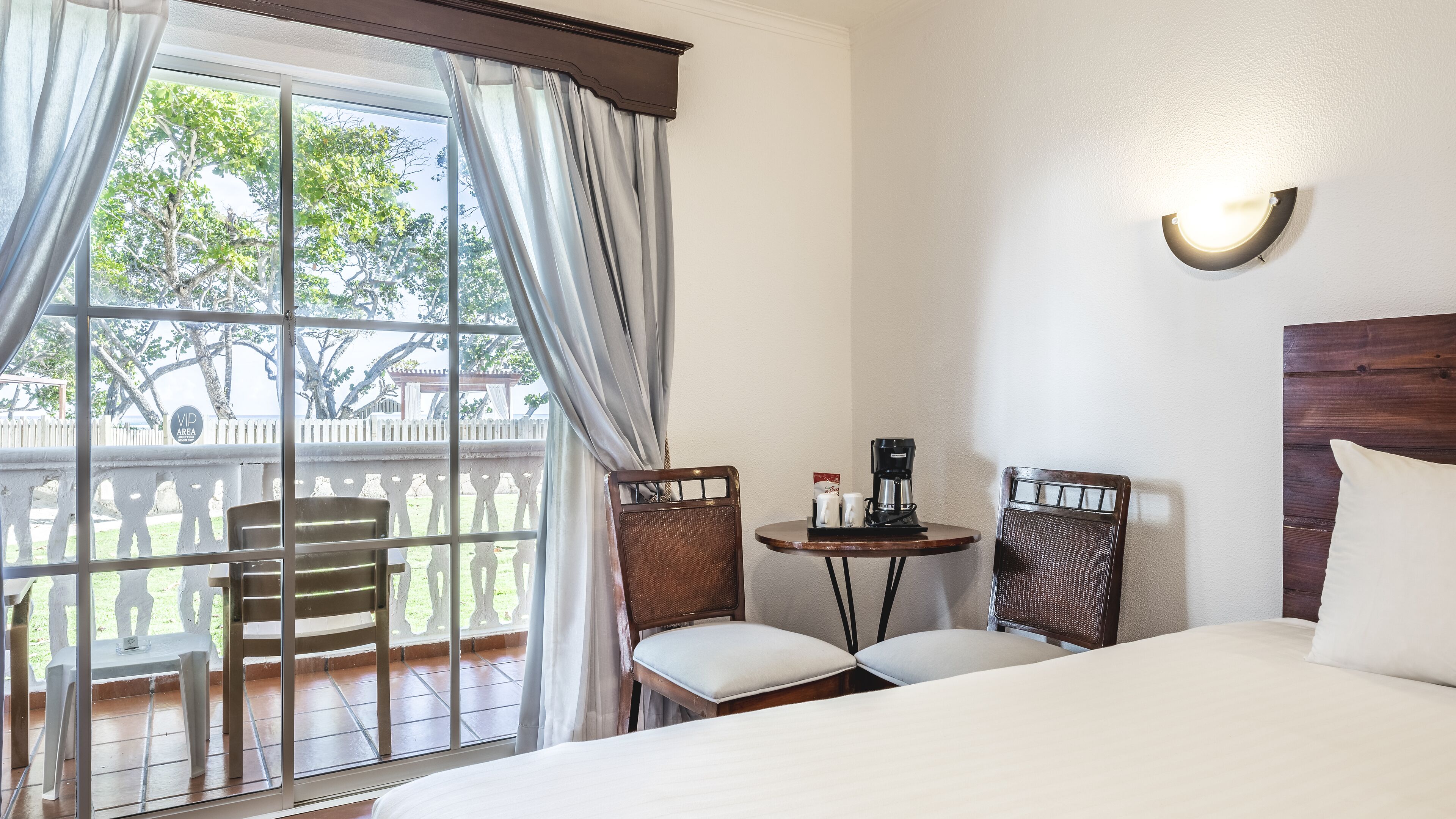 A hotel room with a bed, two chairs, a small table with a coffee maker, and a view of a garden through a window.