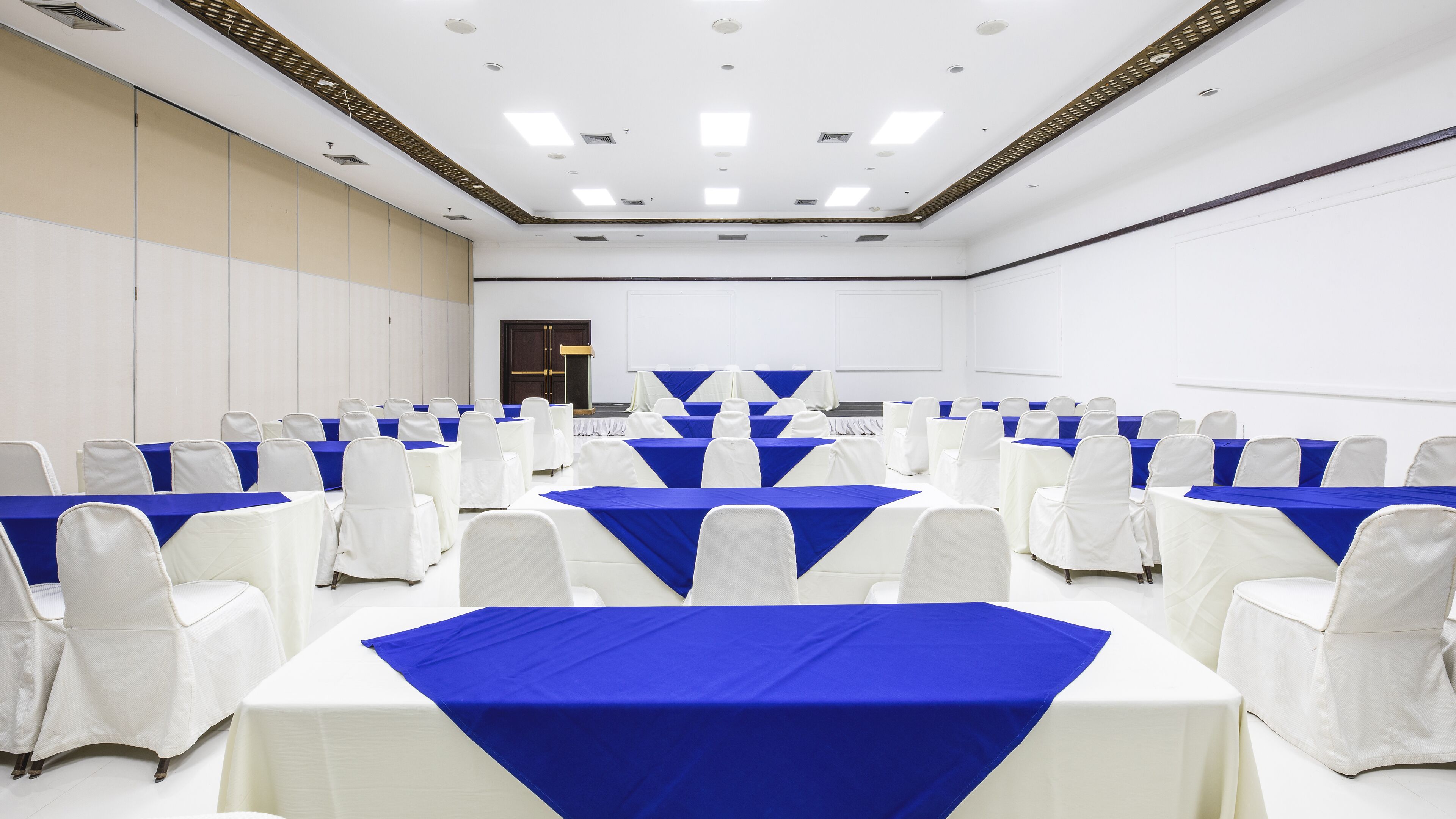 A conference room with rows of tables and chairs, white and blue tablecloths, and a white ceiling with recessed lighting.