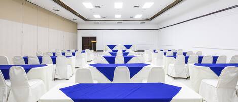 A conference room with rows of tables and chairs, white and blue tablecloths, and a white ceiling with recessed lighting.